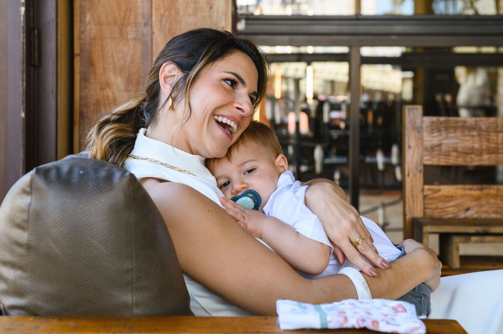Laughing mother holding her baby with a pacifier at an outdoor restaurant table.
