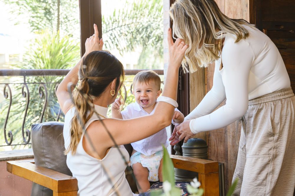 Two women playing and laughing with a happy toddler boy on a wooden bench in a cozy indoor setting.