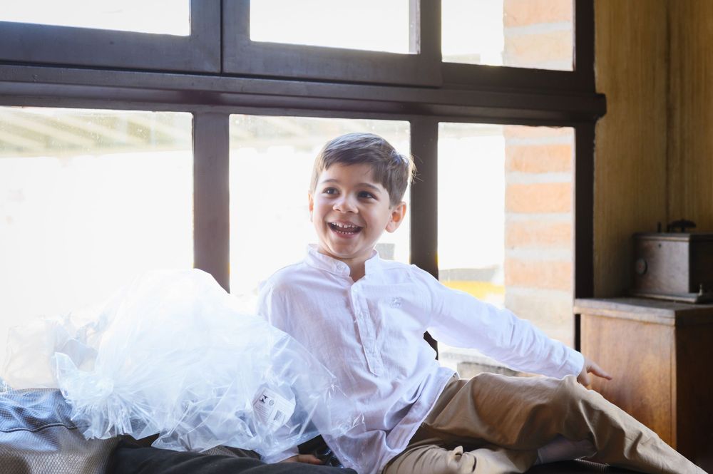 Smiling boy in white shirt sitting near window with white tulle fabric, cheerful expression indoors.