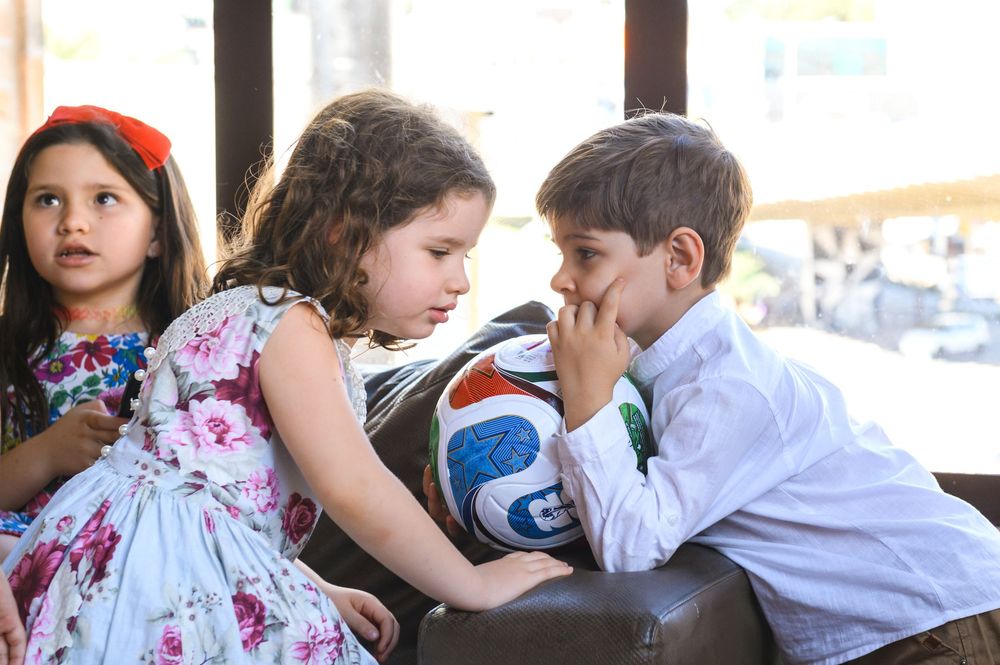 Three young children sitting together on a leather couch, with a soccer ball, dressed in formal attire.