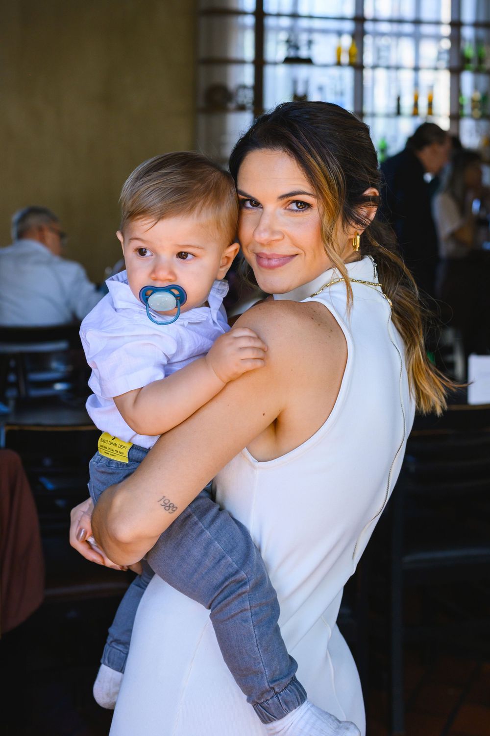 A smiling woman in a white dress holds a baby boy with a pacifier in a warmly lit restaurant setting.