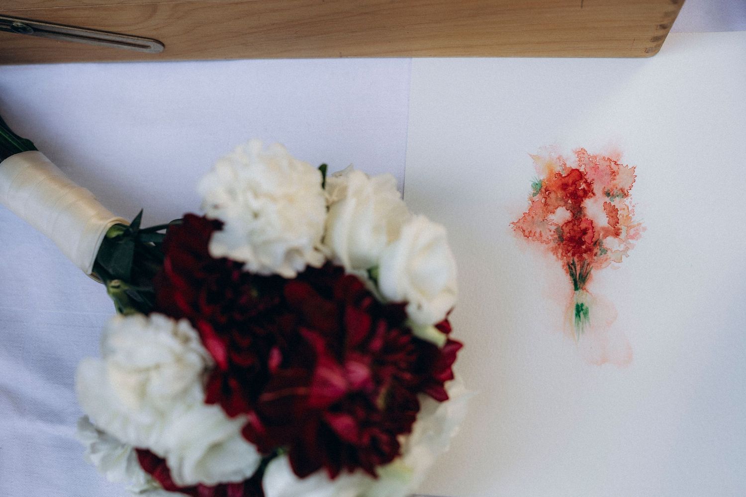 Red and white wedding bouquet with carnations beside a small red floral boutonniere on a white surface.