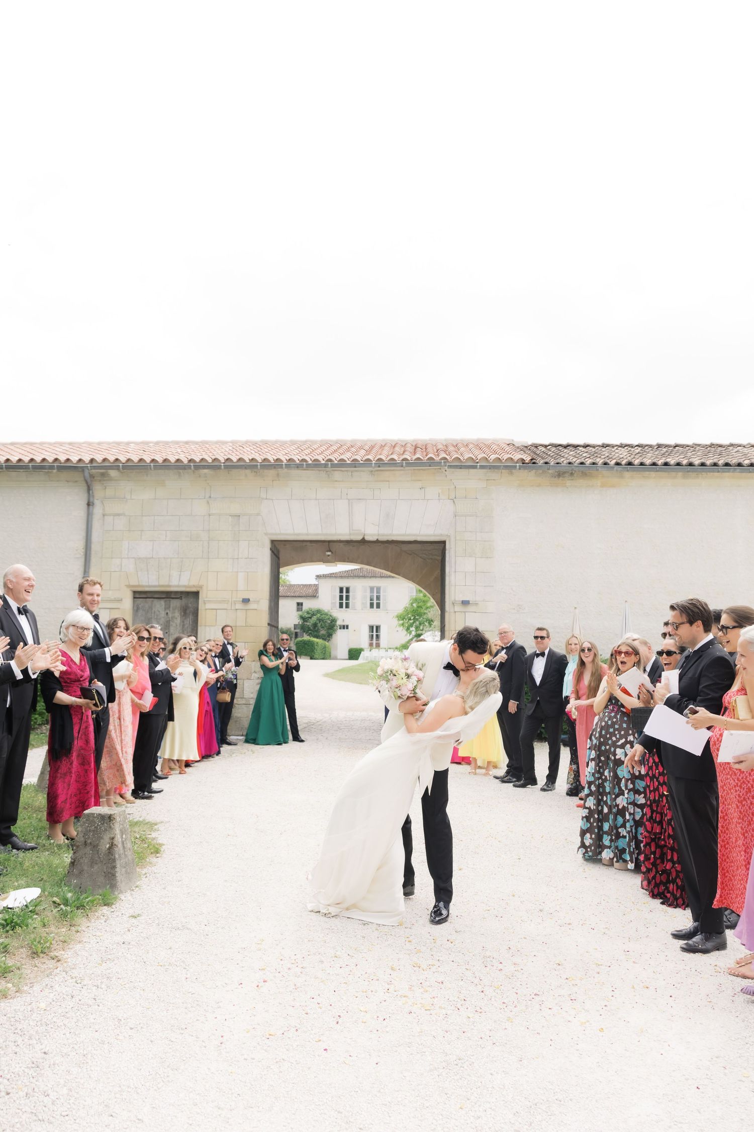 Bride and groom share a romantic dip kiss while wedding guests cheer along a gravel path at an elegant French chateau.