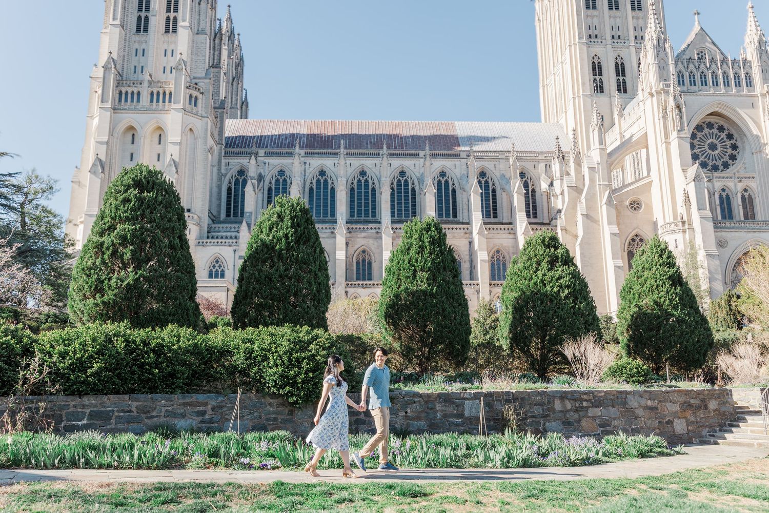Couple walking hand in hand in front of Washington National Cathedral with lush green gardens on a sunny day.