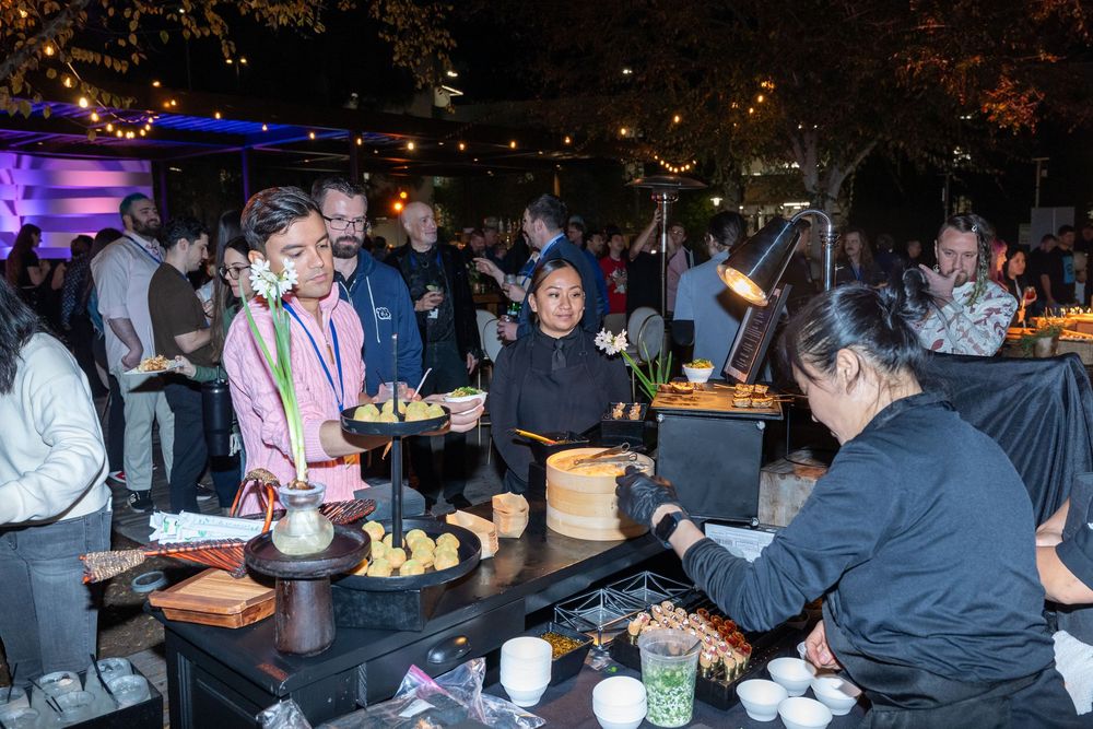 Guests enjoying an outdoor evening food station event with chefs serving dishes under warm lights.