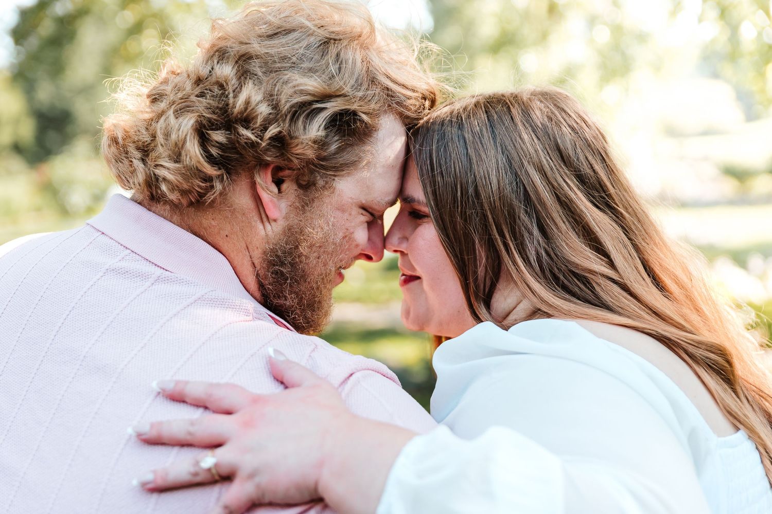 A couple sharing an intimate forehead touch while embracing outdoors, surrounded by lush greenery.