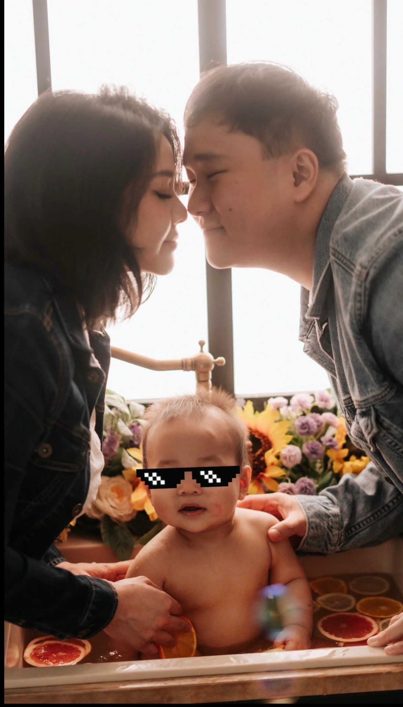 Parents leaning in for a kiss over their baby sitting in a decorative bath with pixel sunglasses overlay.