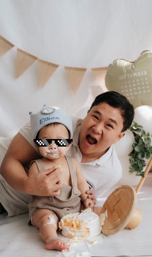 A man joyfully celebrates his baby's first birthday cake smash, with the infant wearing a personalized hat and pixelated sunglasses.
