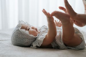 A newborn baby in a floral outfit lies on a soft blanket, reaching tiny hands toward an adult's hand above.