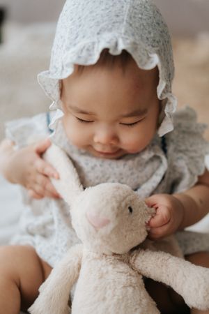 A smiling baby in a floral bonnet and dress gently holds a soft plush bunny toy.