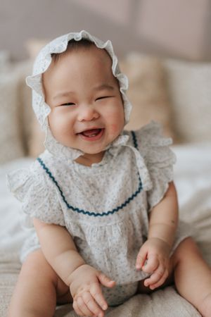 Smiling baby girl in floral romper and white lace bonnet sitting happily on a bed.