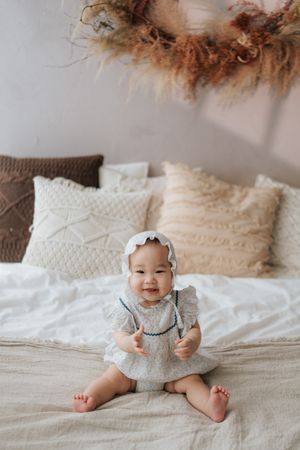 Smiling Asian baby girl in white lace bonnet and dress sitting on a bed with neutral boho pillows.