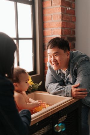 Smiling Asian father leaning over baby bathtub with infant while mother watches near brick wall window.