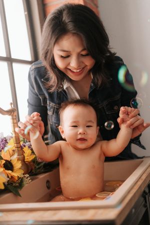 Smiling Asian mother helping her baby stand in a small bathtub with sunflowers and soap bubbles nearby.