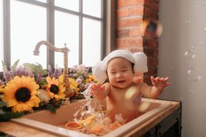 Happy baby in white hat playing in flower-filled bathtub with bubbles floating nearby.