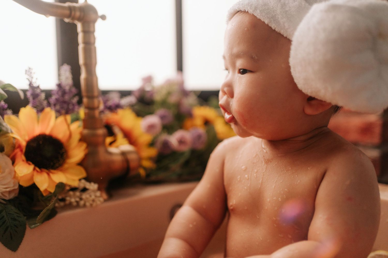 Cute Asian baby in a bathtub with a white towel on head, surrounded by sunflowers and colorful blooms.