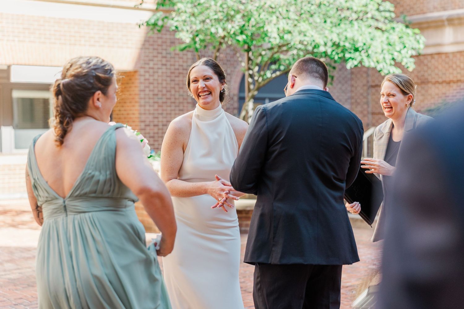 Bride in white halter gown laughs joyfully while holding hands with groom outdoors at wedding ceremony.