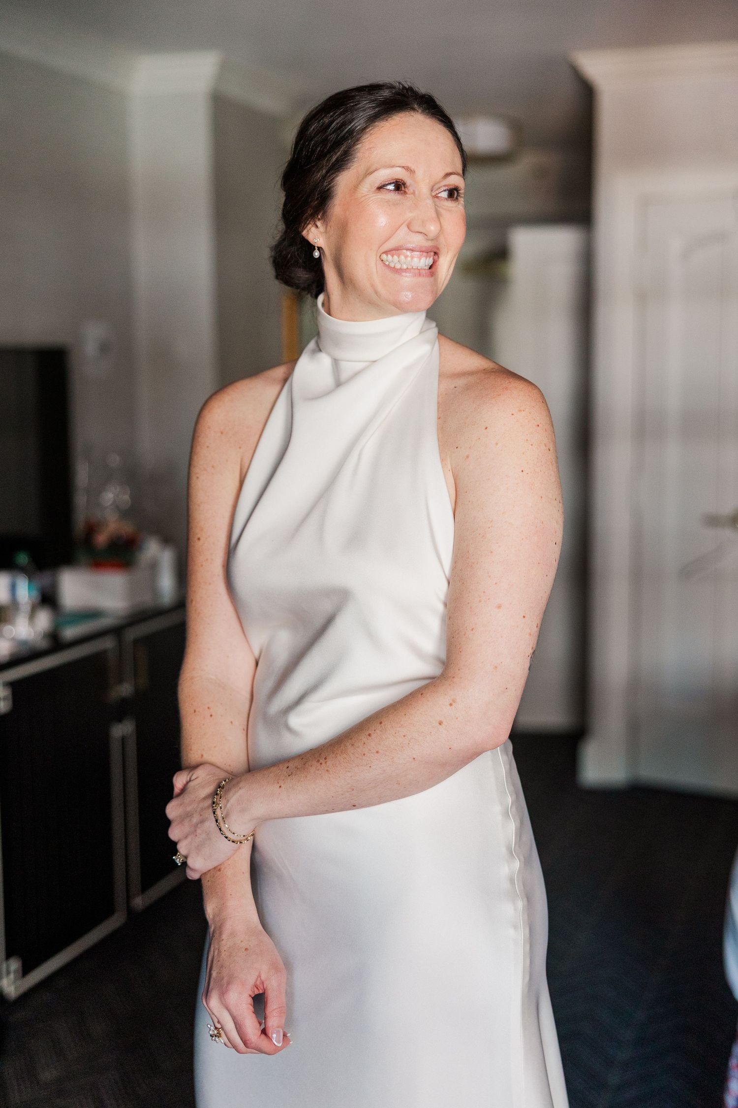 Smiling bride in elegant white halter neck gown poses indoors, hair styled in a chic updo with pearl earrings.