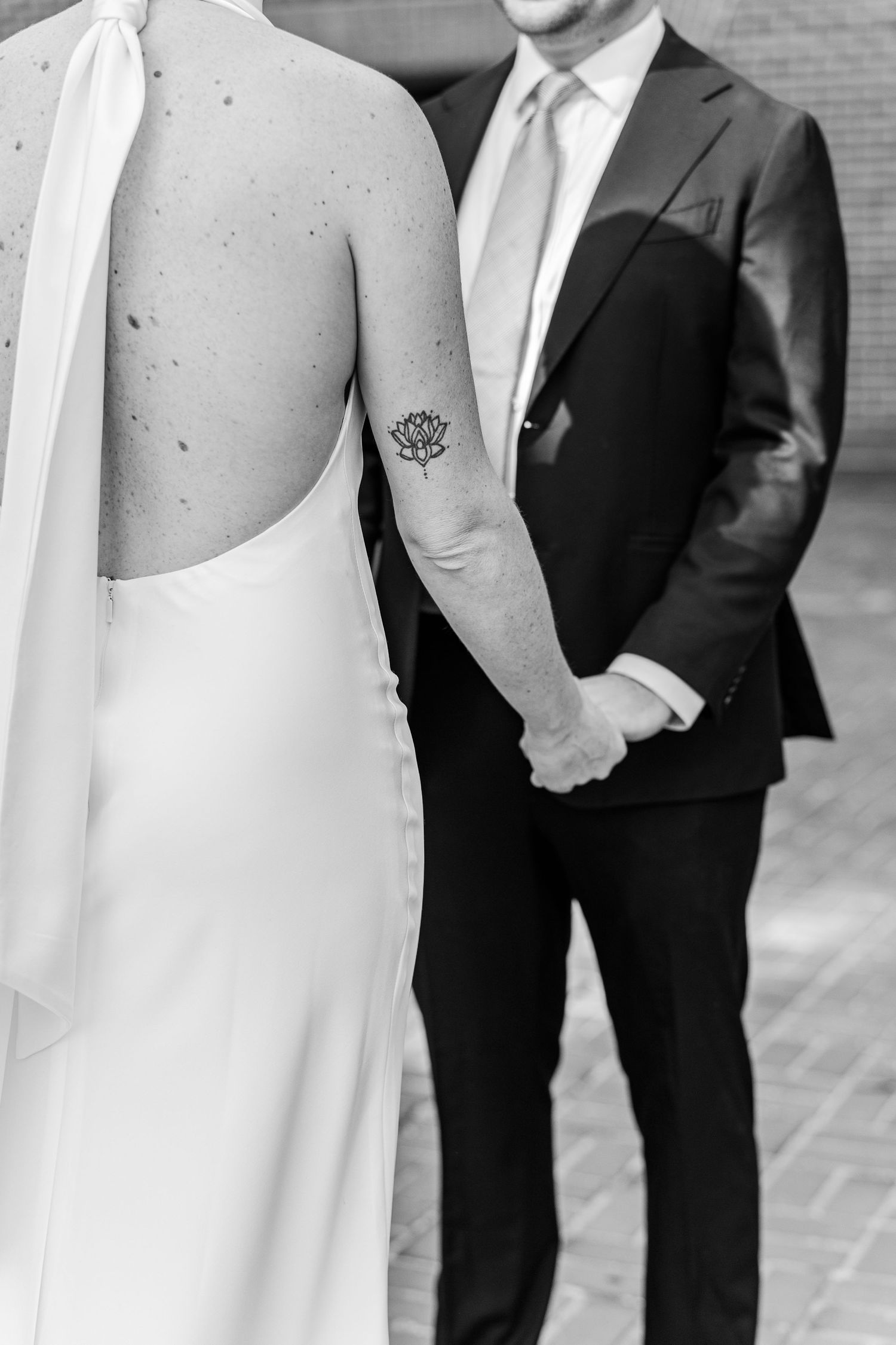 Black and white photo of bride with lotus tattoo holding hands with groom in suit at wedding.