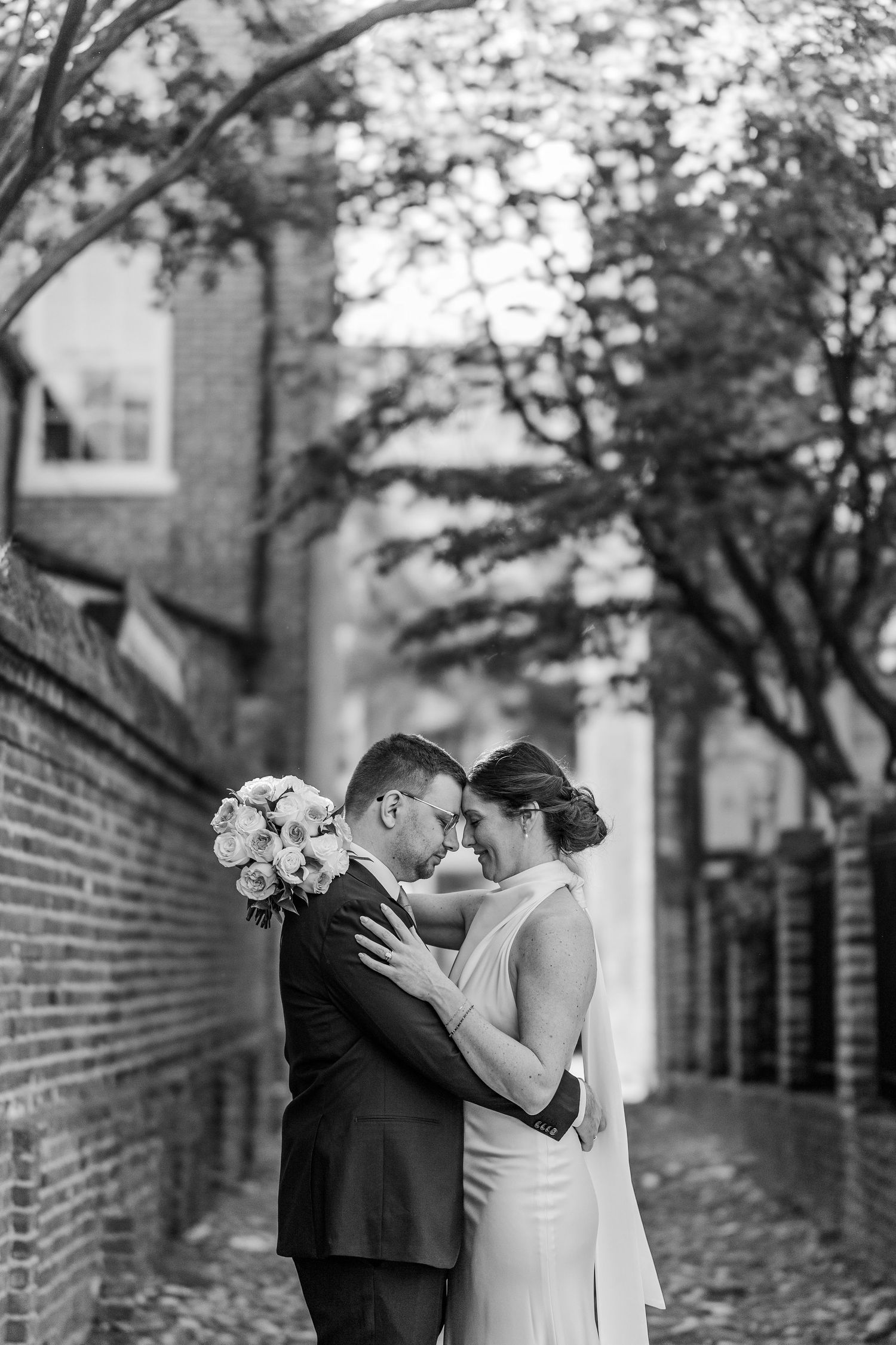Black and white photo of a bride and groom embracing in a charming brick alleyway, sharing an intimate moment on their wedding day.