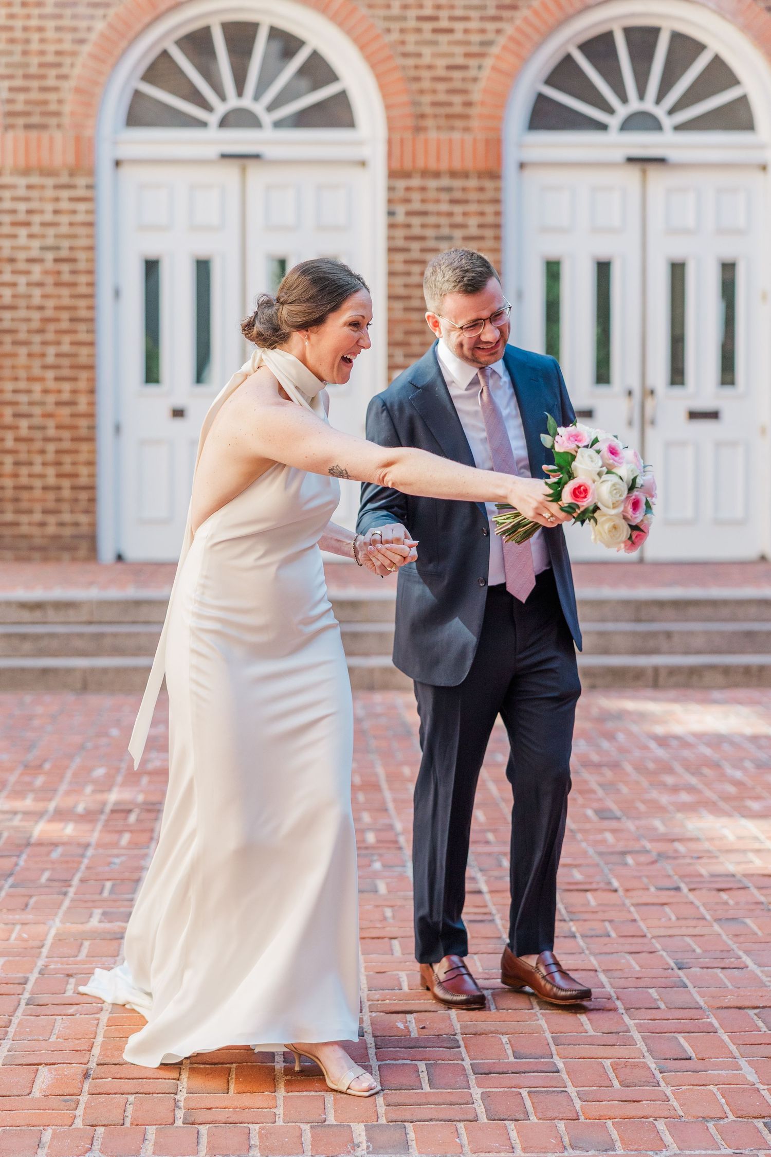 Bride in white gown holds bouquet while laughing with groom in navy suit on brick steps outside church.