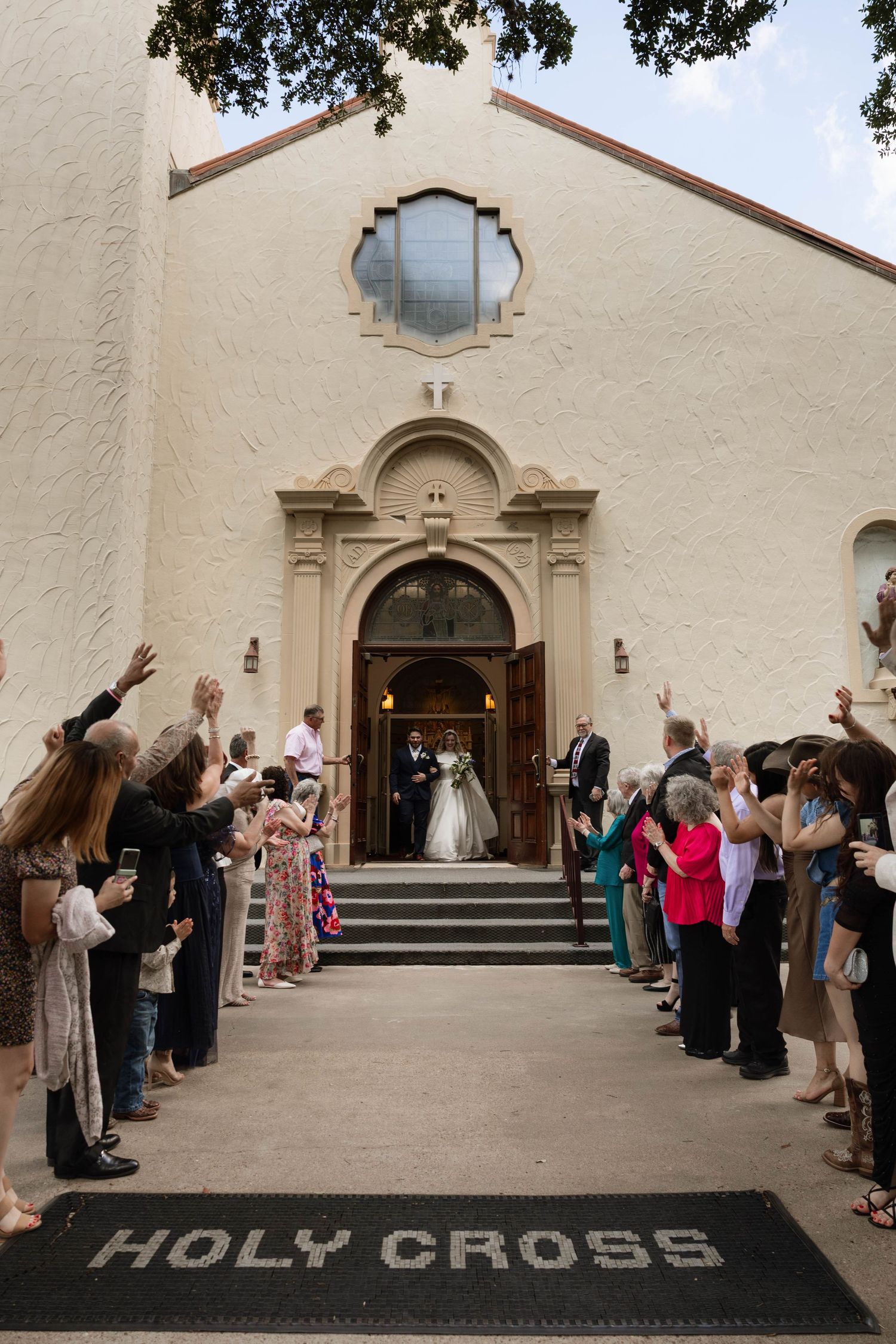 Newlyweds exiting Holy Cross church as guests celebrate with raised hands lining the steps after the ceremony.