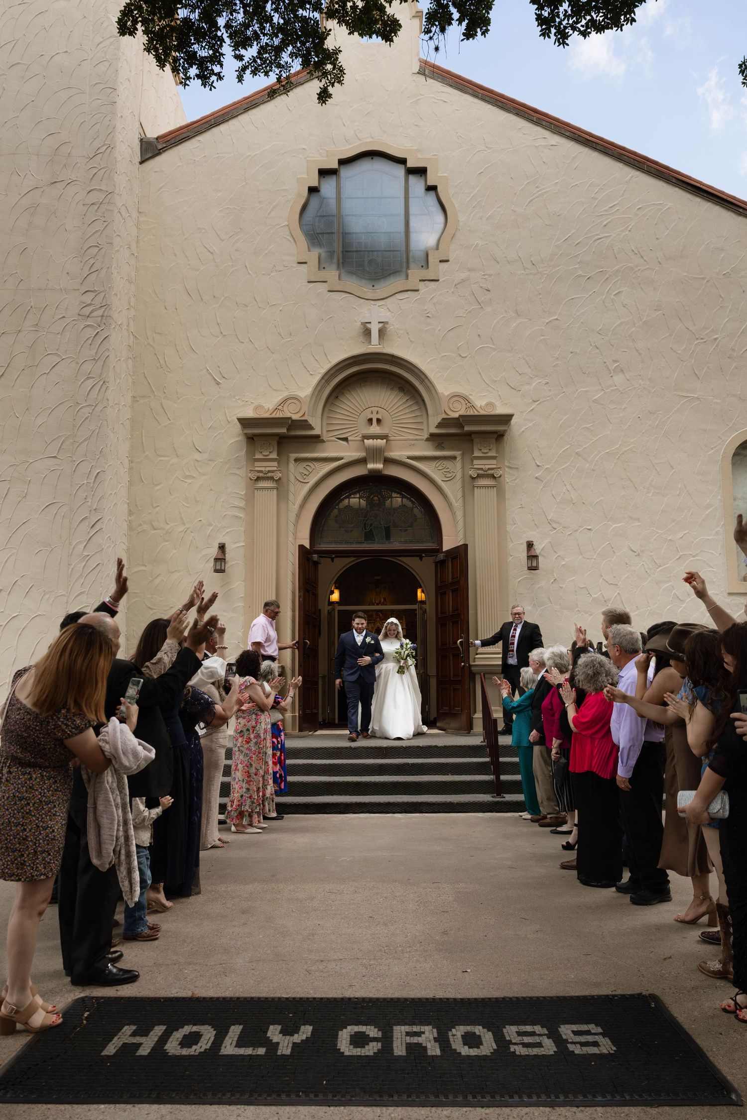 Newlyweds exiting Holy Cross church as guests celebrate with raised hands lining the steps after the ceremony.
