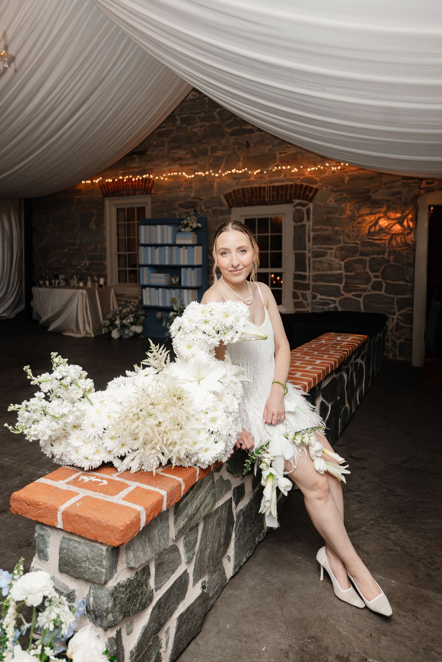 Bride in short white dress sits on brick ledge holding large white floral bouquet at stone venue.
