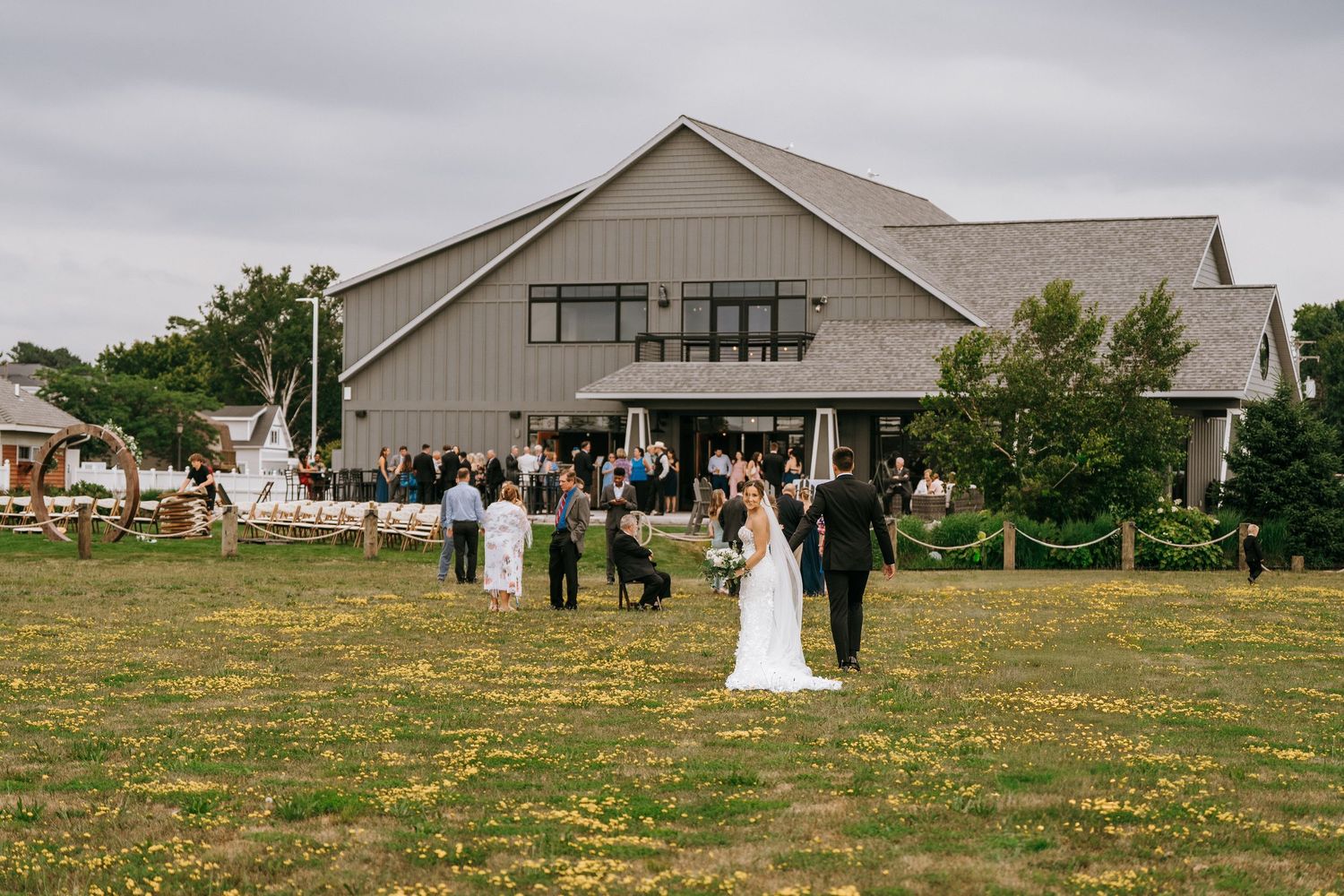 Outdoor wedding ceremony on a green field with guests gathered near a large gray barn venue under a cloudy sky.