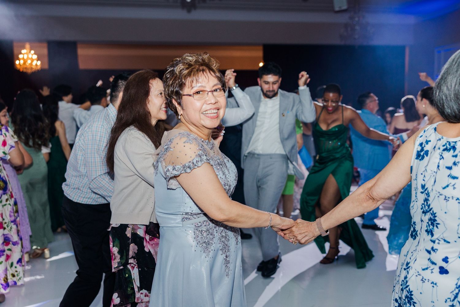 Guests dancing joyfully at a wedding reception, wearing formal attire in shades of blue, green, and floral patterns.