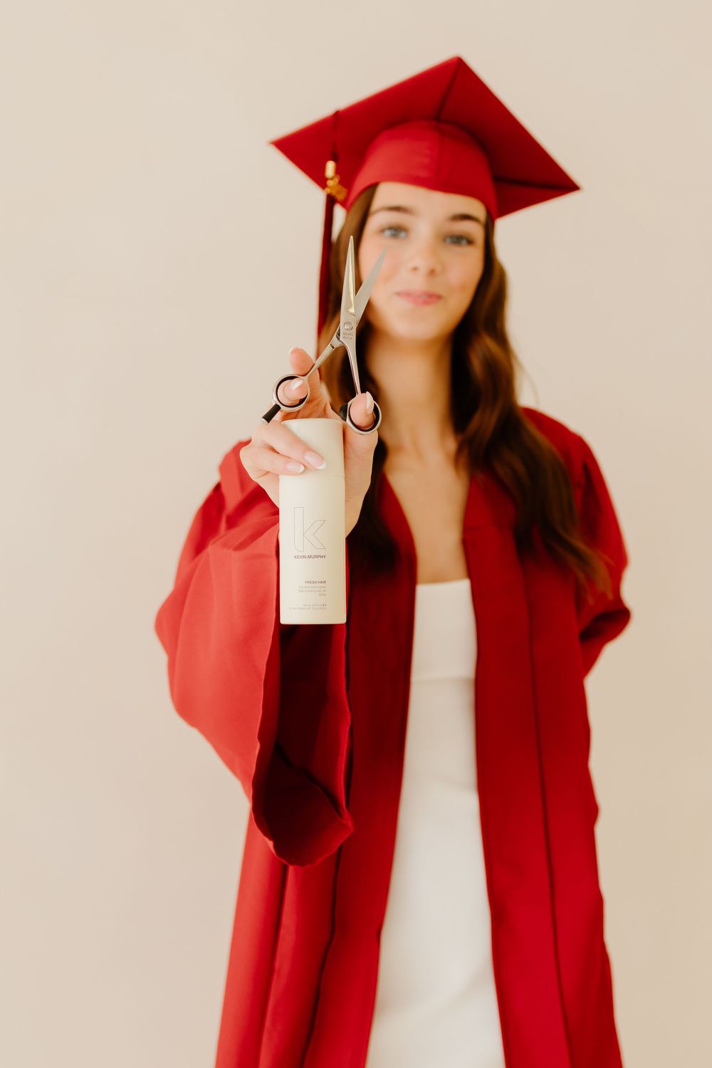 Young woman in red graduation gown extending scissors and product toward camera with soft focus.