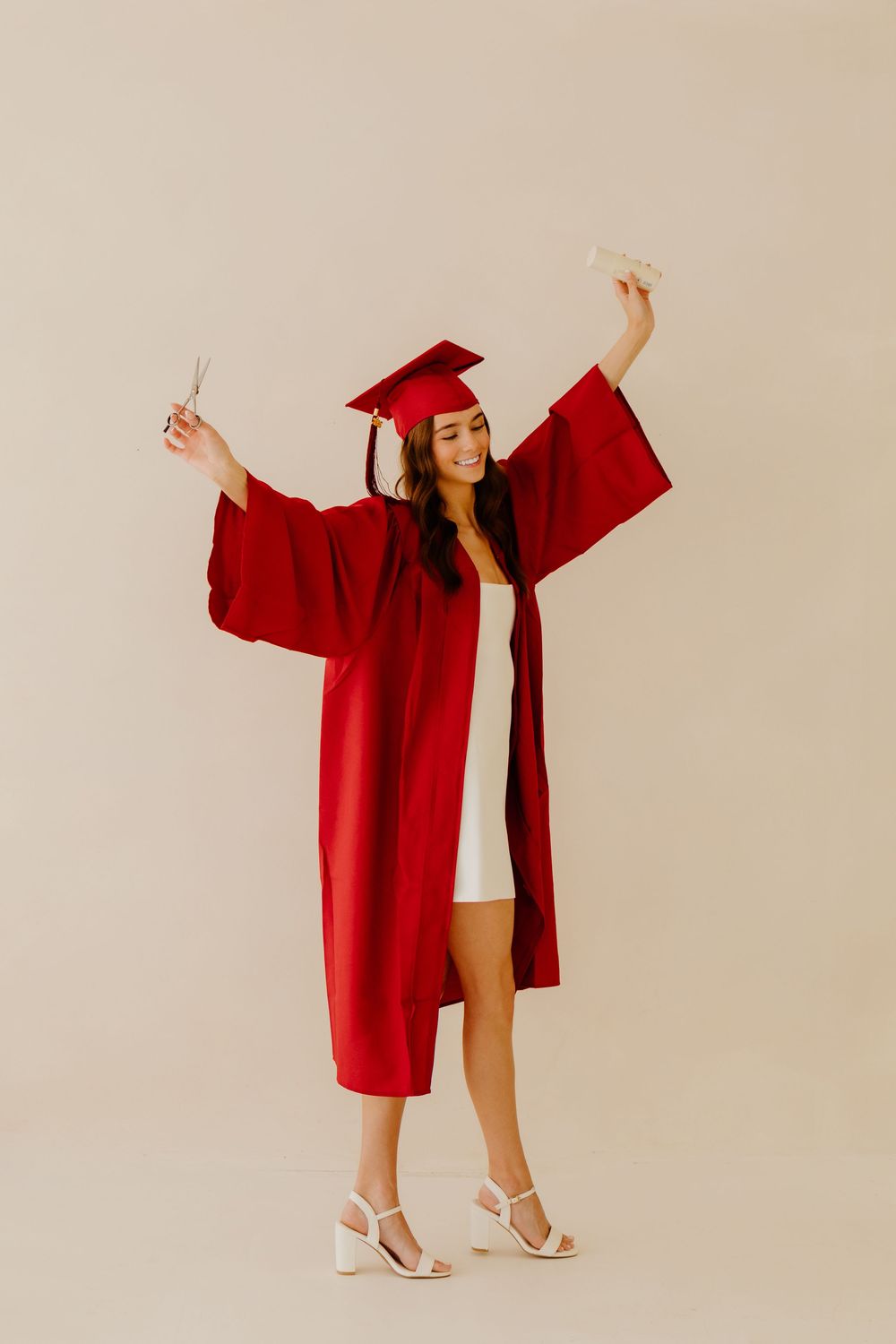 Joyful graduate in red cap and gown celebrating with arms raised holding scissors and diploma.