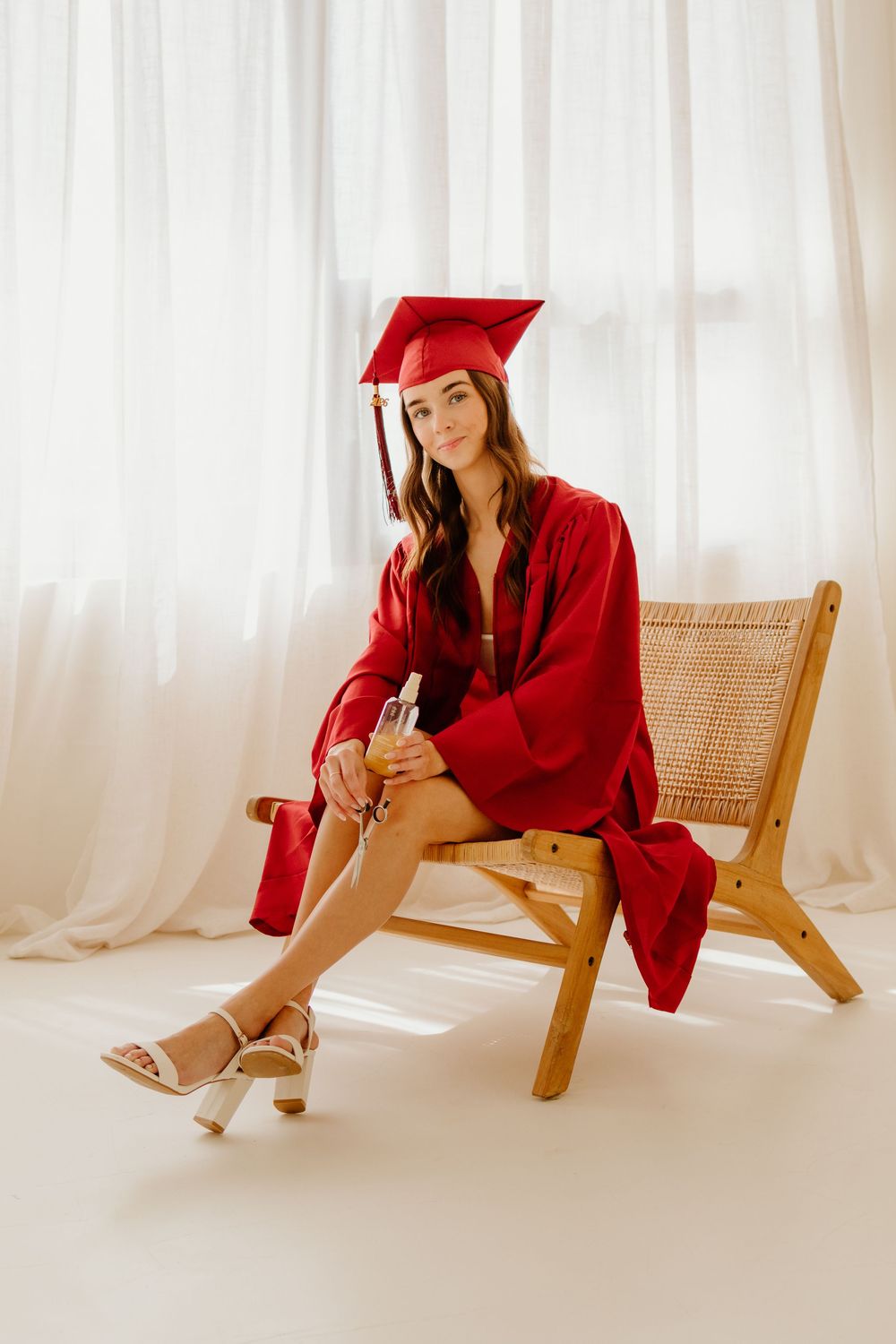 Young woman in red graduation cap and gown sitting elegantly on a wooden chair holding a diploma.