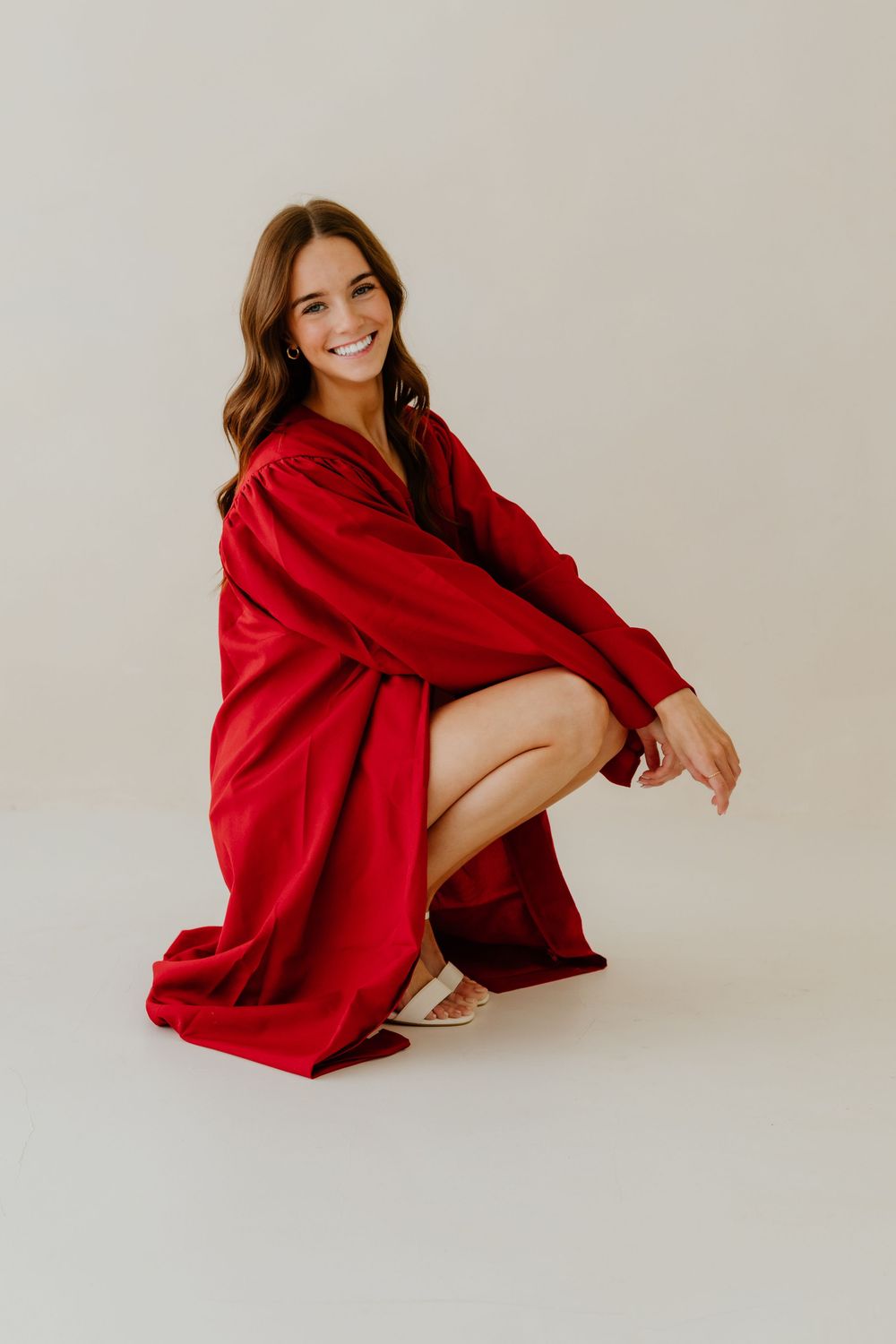 Cheerful woman in red graduation gown crouching and smiling against light neutral background.