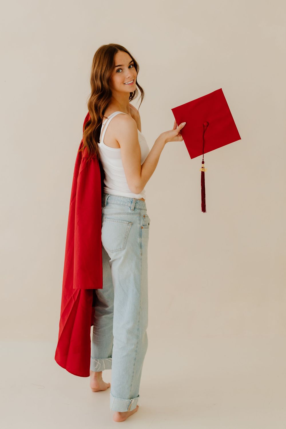 Young woman in white tank and jeans draping red graduation gown over shoulder holding cap.