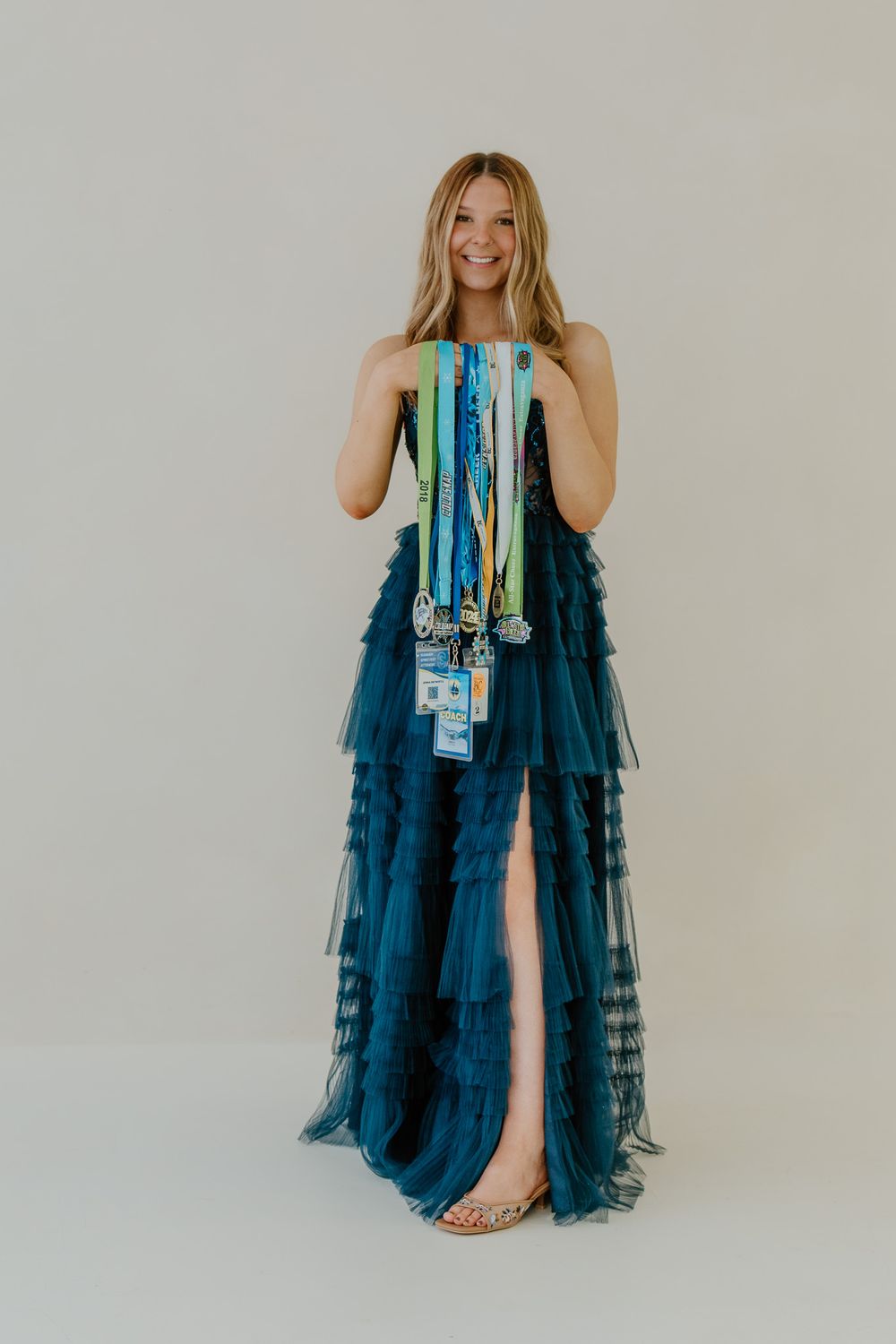 Smiling blonde woman in elegant teal tulle gown holding cheerleading medals and competition badges.