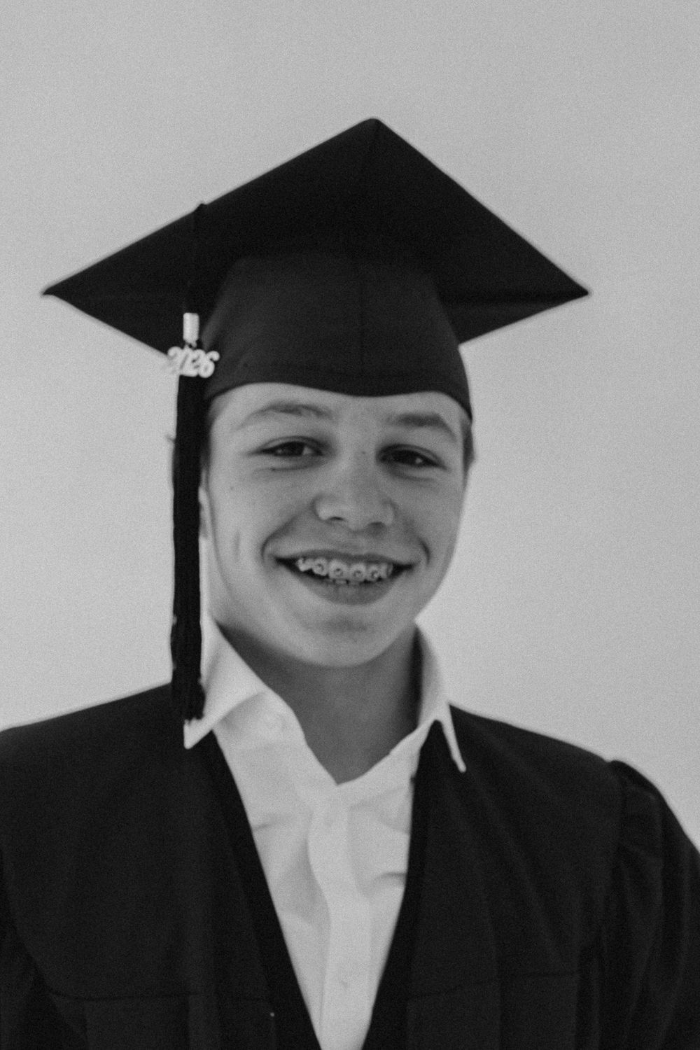 Smiling teen in black graduation cap and gown wearing braces in black and white portrait photo.