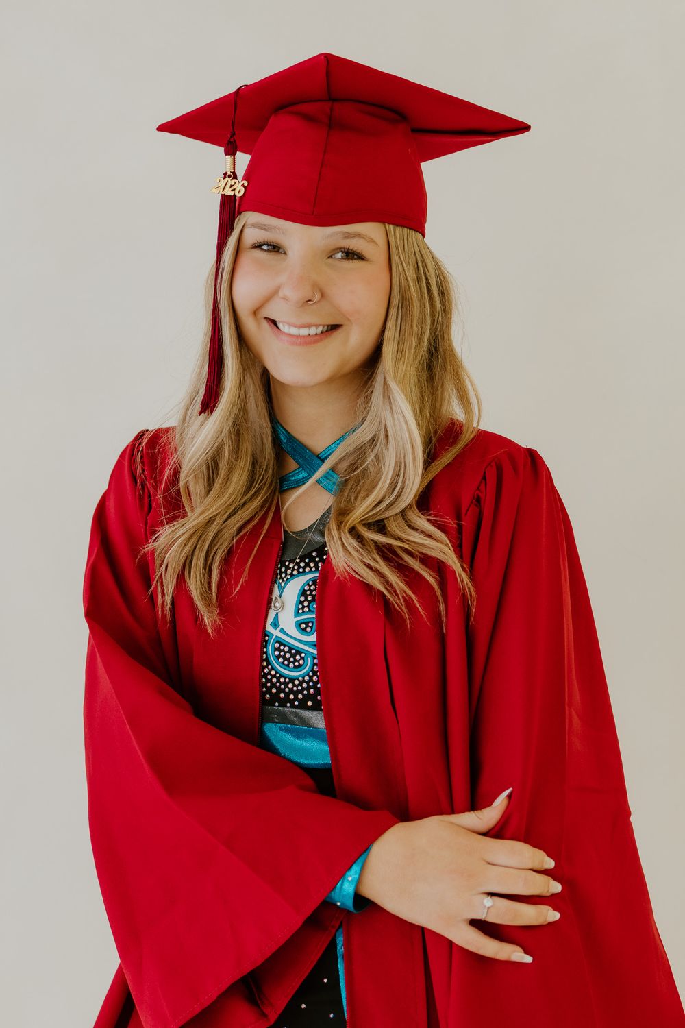 Smiling graduate in red cap and gown wearing a cheerleading uniform underneath, posing with arms crossed.