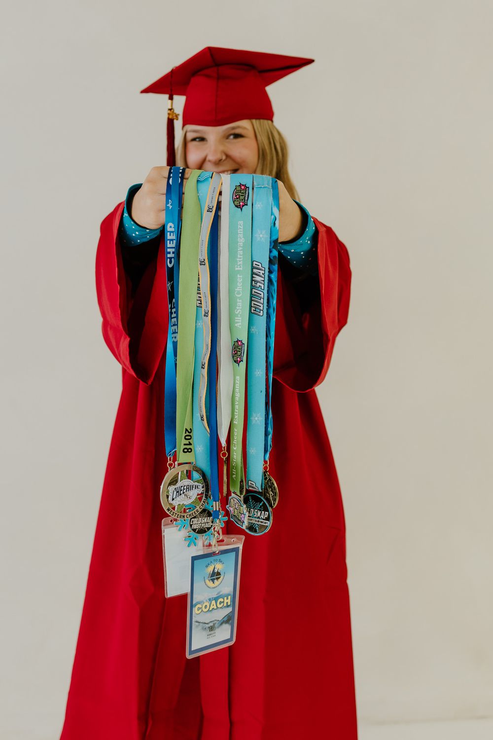 Smiling graduate in red cap and gown holding up cheerleading medals and colorful award ribbons.