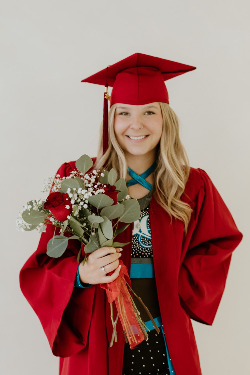 Cheerful graduate in red cap and gown holding a bouquet of red roses and eucalyptus.