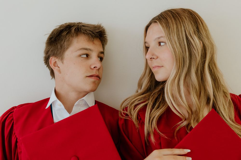 Two teens in red graduation gowns looking at each other closely against a white wall.