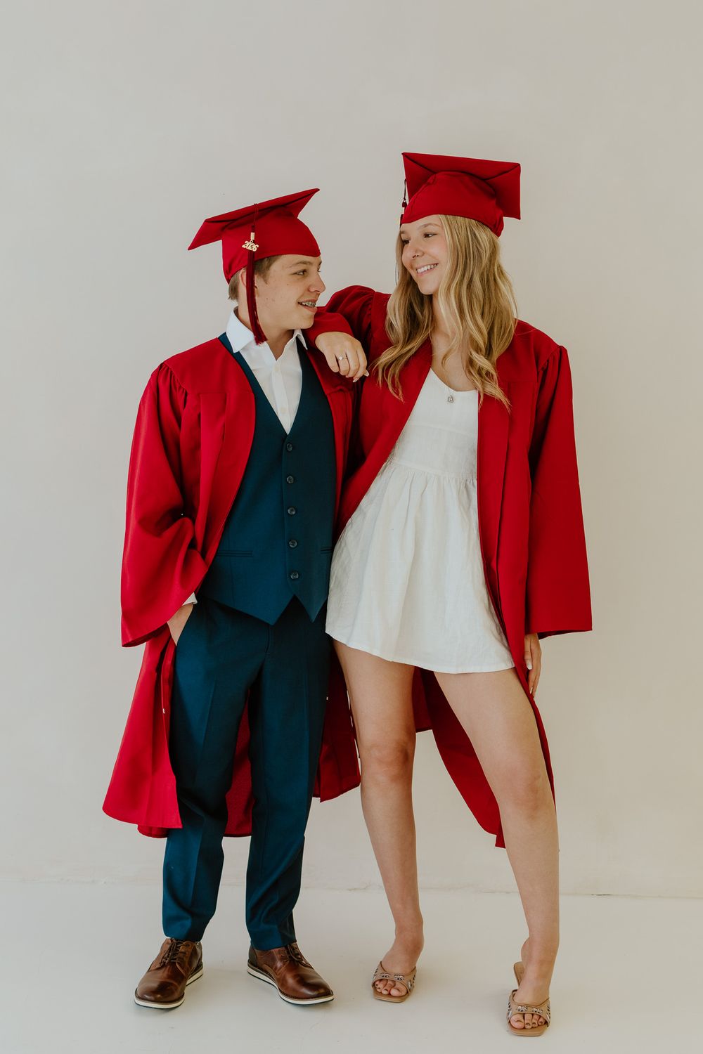 Two smiling teens in red graduation caps and gowns standing together in a full-length portrait.