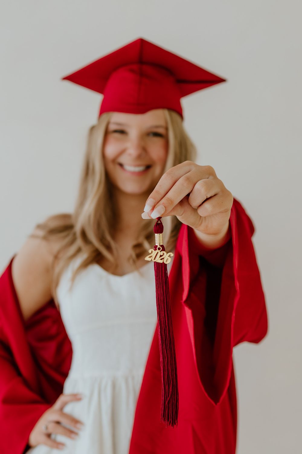 Smiling blonde graduate in red cap and gown proudly displays a burgundy 2026 graduation tassel.