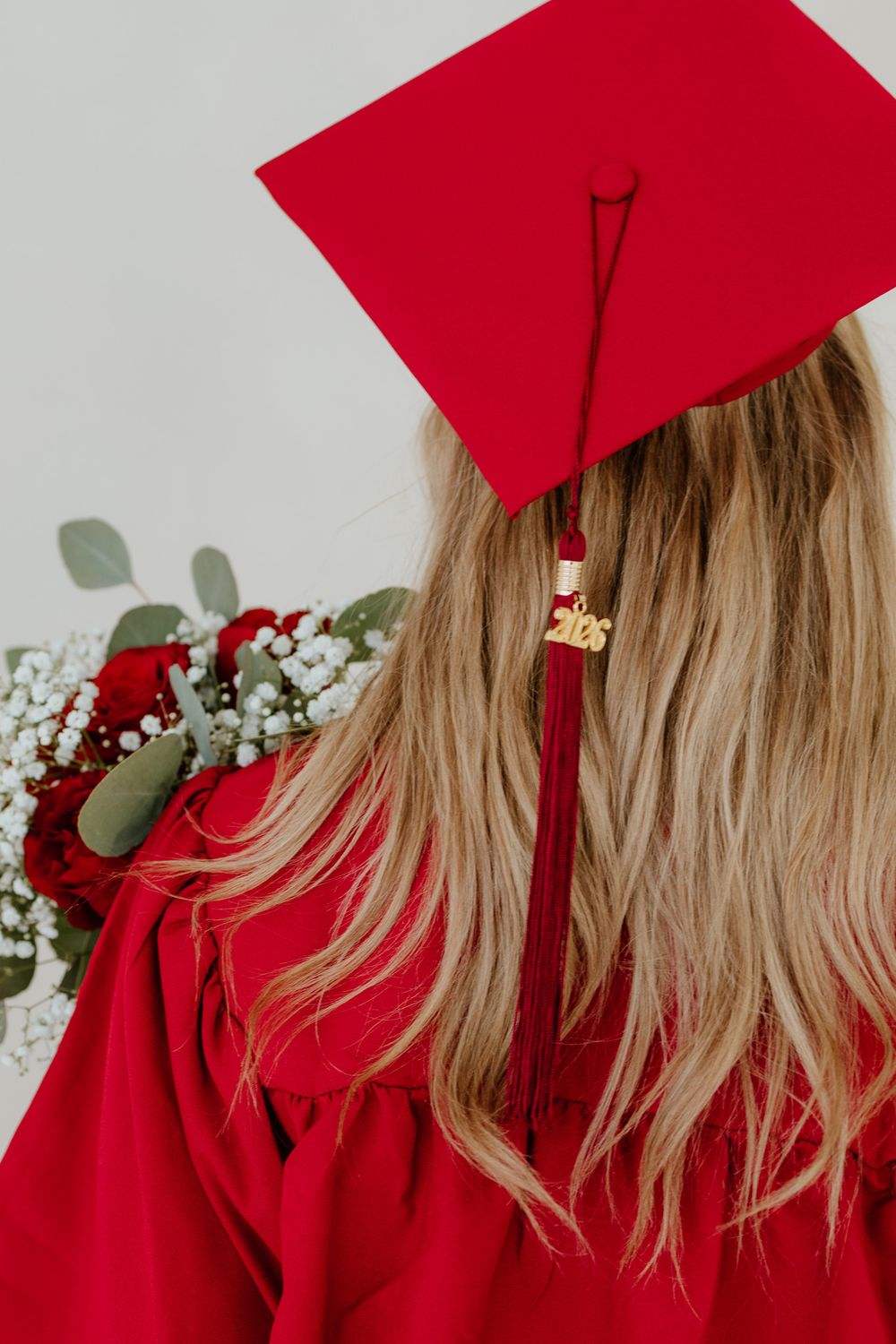 Back view of a blonde graduate in red cap and gown with a 2026 tassel and red floral bouquet.