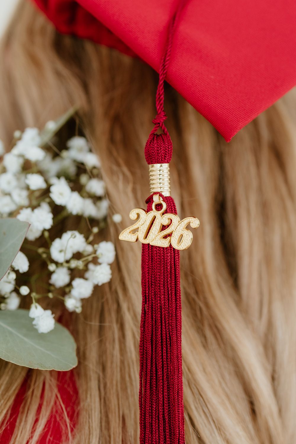Close-up of a gold 2026 graduation tassel charm resting in blonde hair beside white flowers.