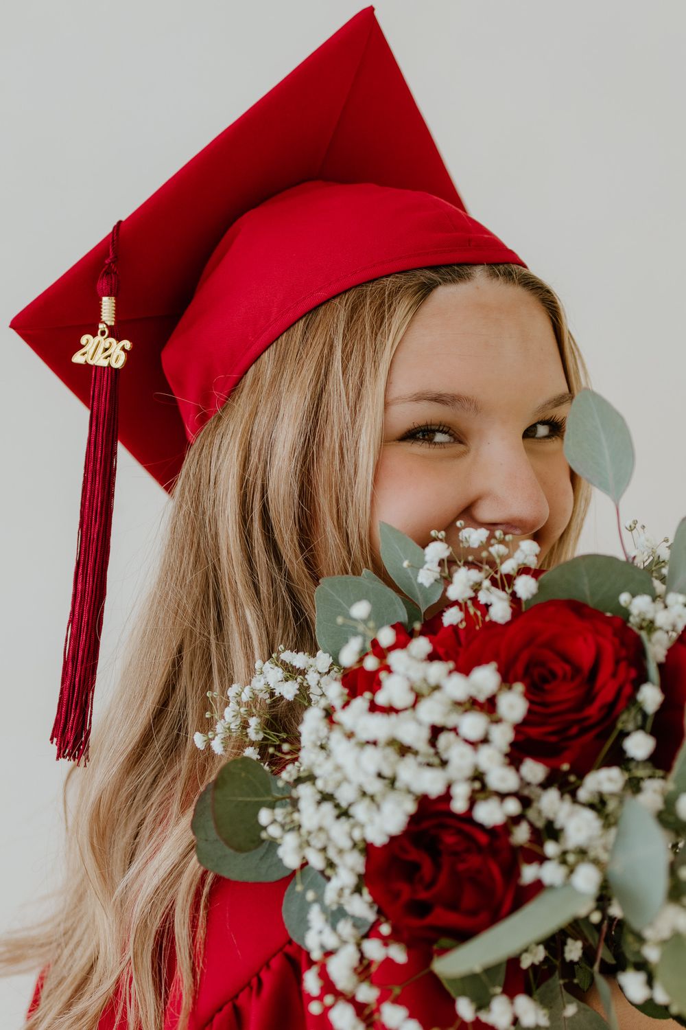 Smiling blonde graduate in red 2026 cap holds red rose bouquet close to her face.