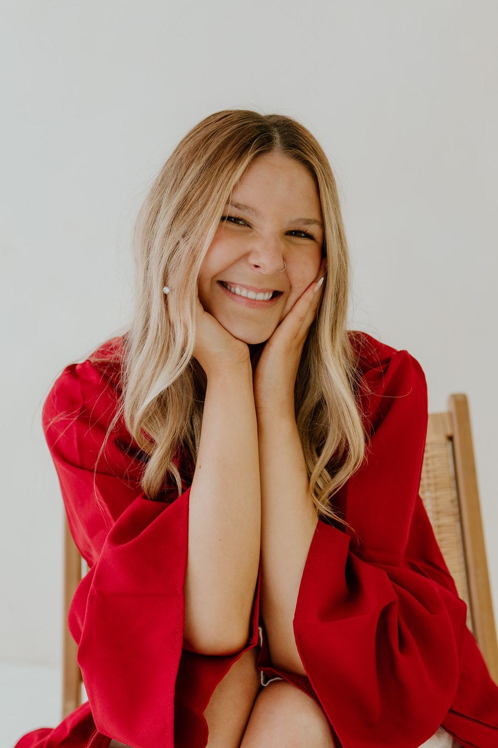Smiling blonde woman in red dress sitting on chair with hands on cheeks.