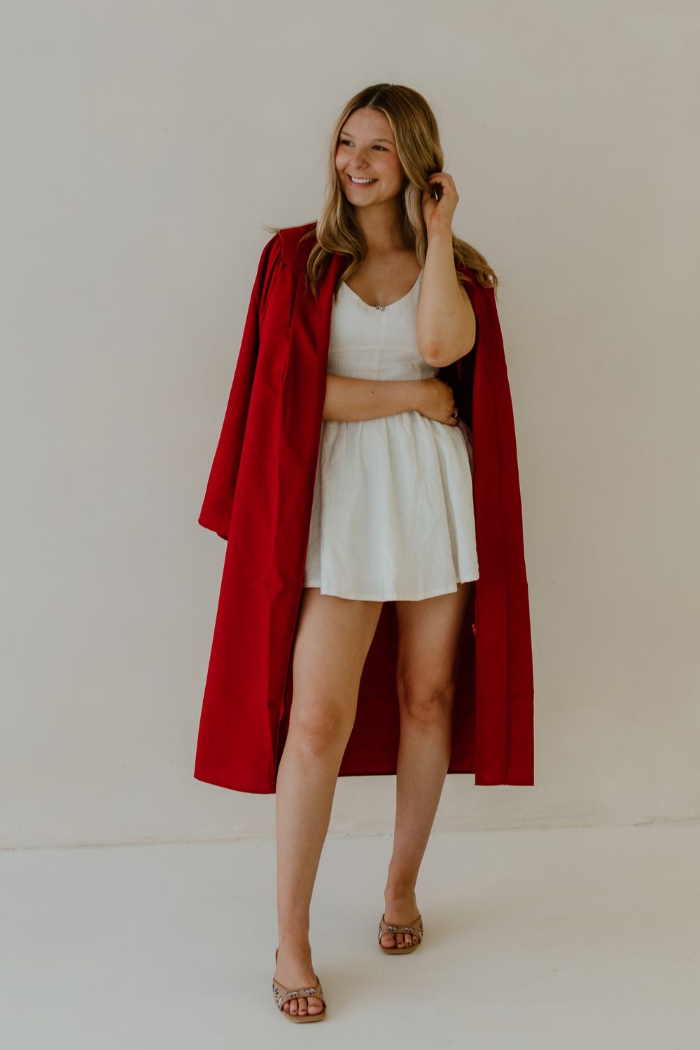 Smiling blonde woman in white dress with open red graduation gown standing.