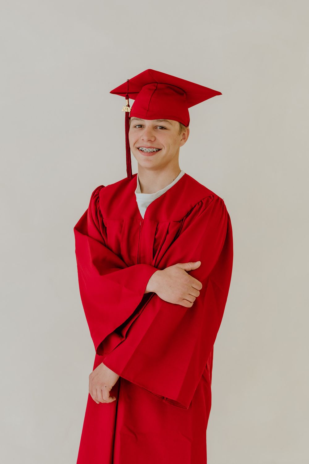 Smiling teen in red graduation cap and gown with arms crossed for senior portrait photo.