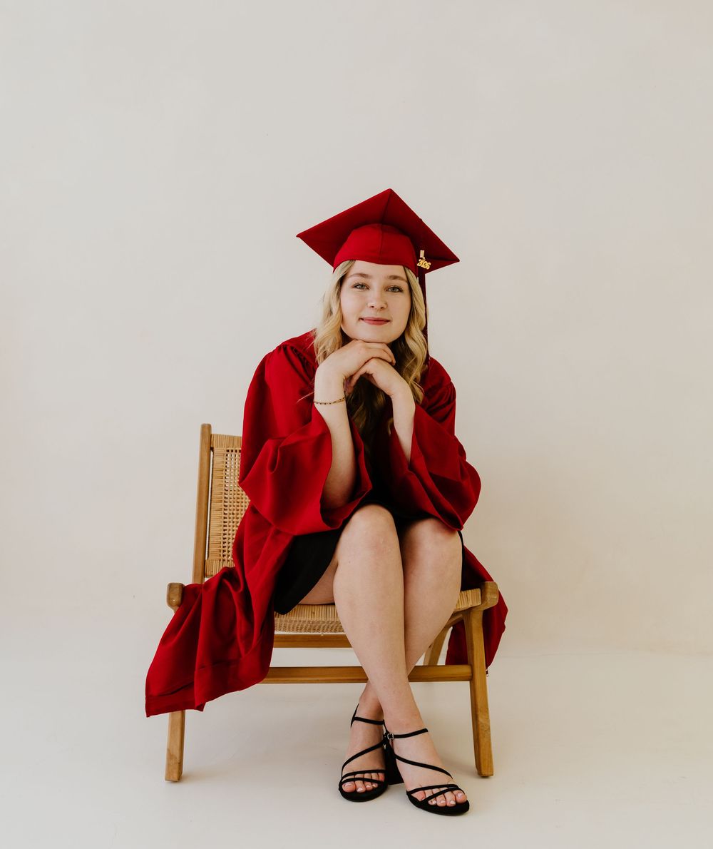 Smiling graduate in red cap and gown seated on wooden chair for senior portrait photo.